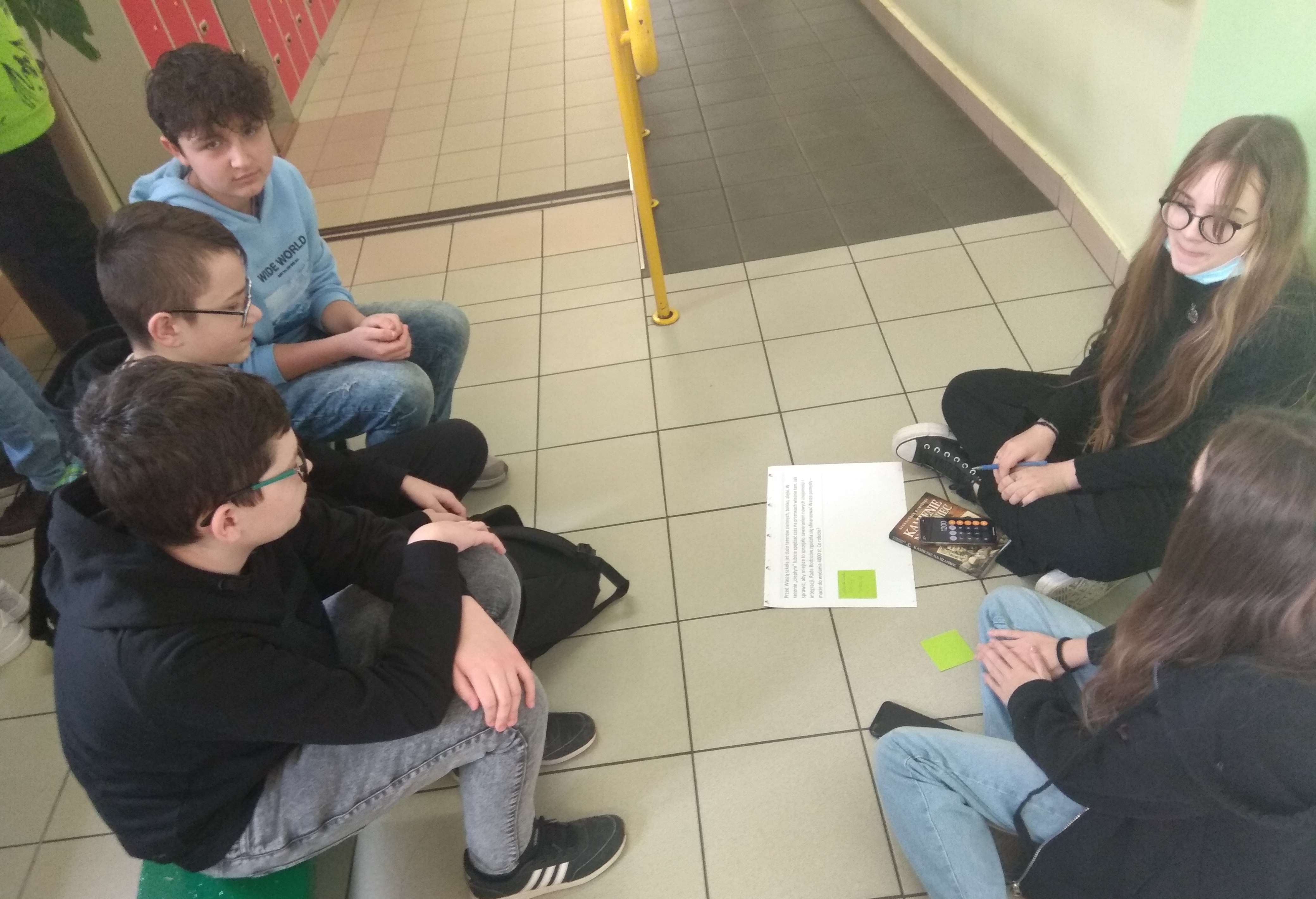 Grupa dzieci siedząca na szkolnym korytarzu, wpisująca pomysły na projekty na kartce